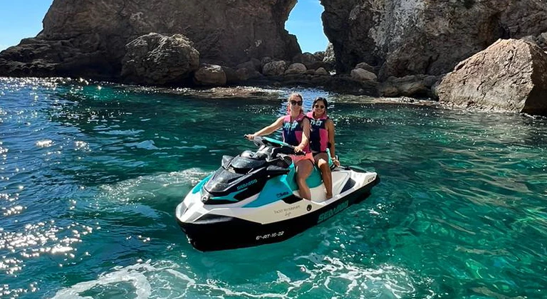 jetski-tour-ibiza-couple-jetskiclub Two women riding a Sea-Doo jet ski during a jet ski tour with jetski.club in Ibiza crystal clear turquoise waters